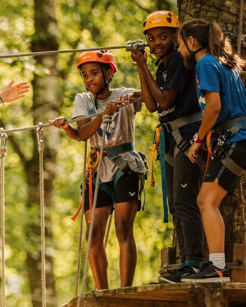 Muddy hands helping a teammate up a wooden obstacle, with the team cheering around them