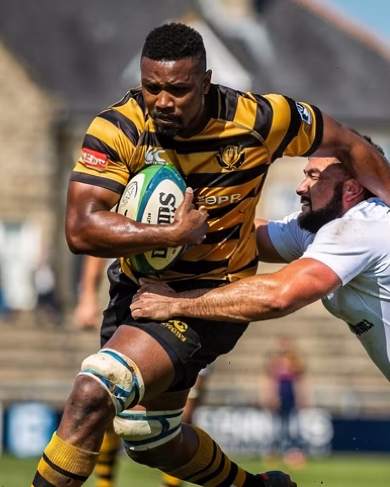 A player being tackled at a European club ground, stone clubhouse in the background