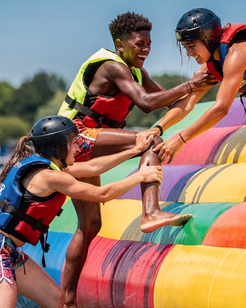 Three teenage teammates working together to climb an inflatable obstacle at a water park