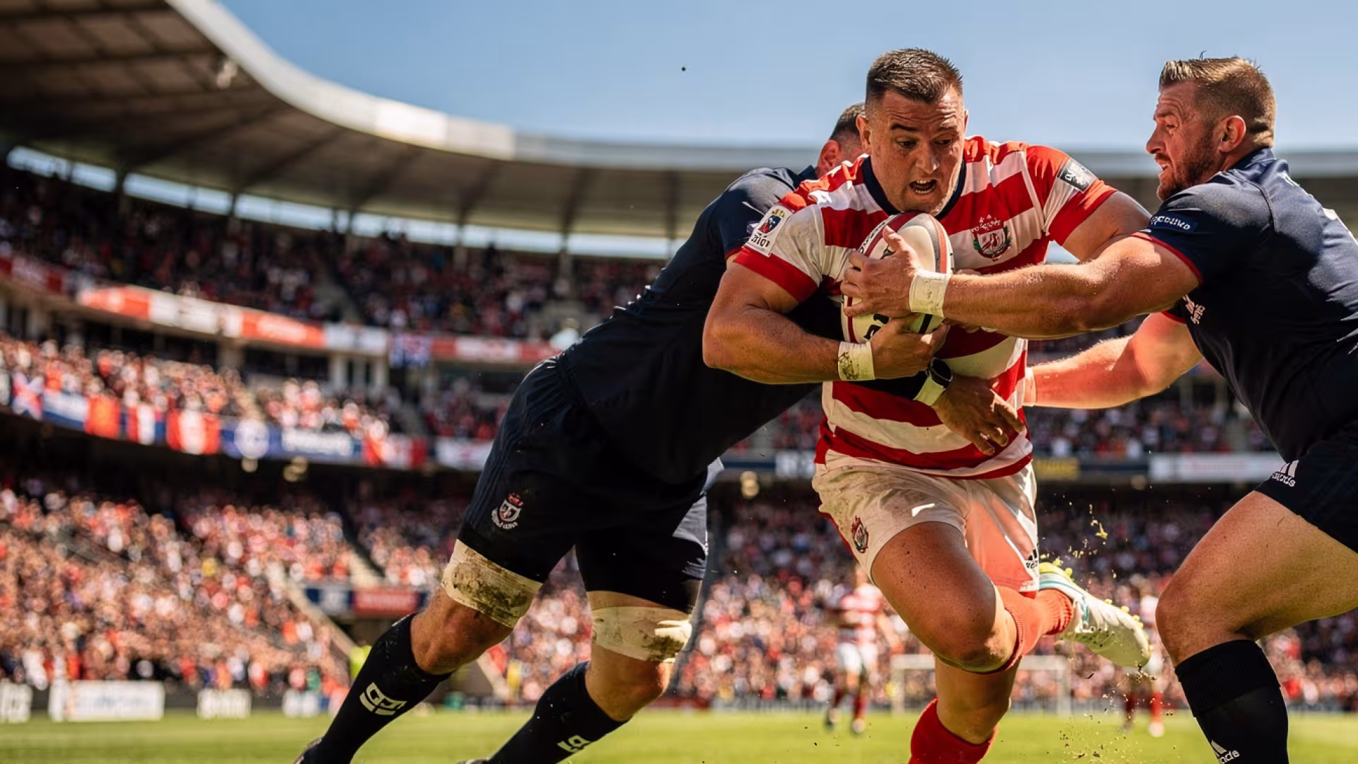 A rugby player powering through two tacklers during a professional match at a packed stadium